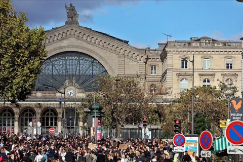 A large crowd of marchers in front of a Paris railway station with an arched glass facade.