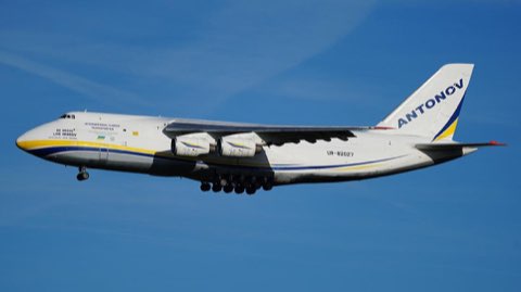 A white Antonov An-124 cargo plane in Ukrainian blue-and-yellow livery flying against a blue sky.