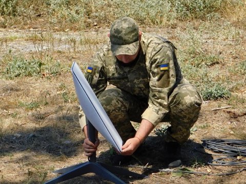 A Ukrainian soldier in camouflage kneels to set up a white Starlink satellite dish on a stand in a dry field.